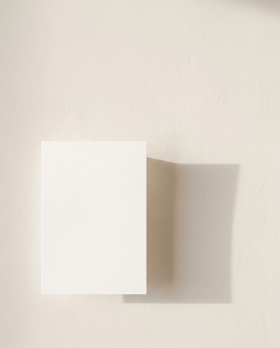 A single sheet of white paper resting on a warm plaster wall, casting a long soft shadow.